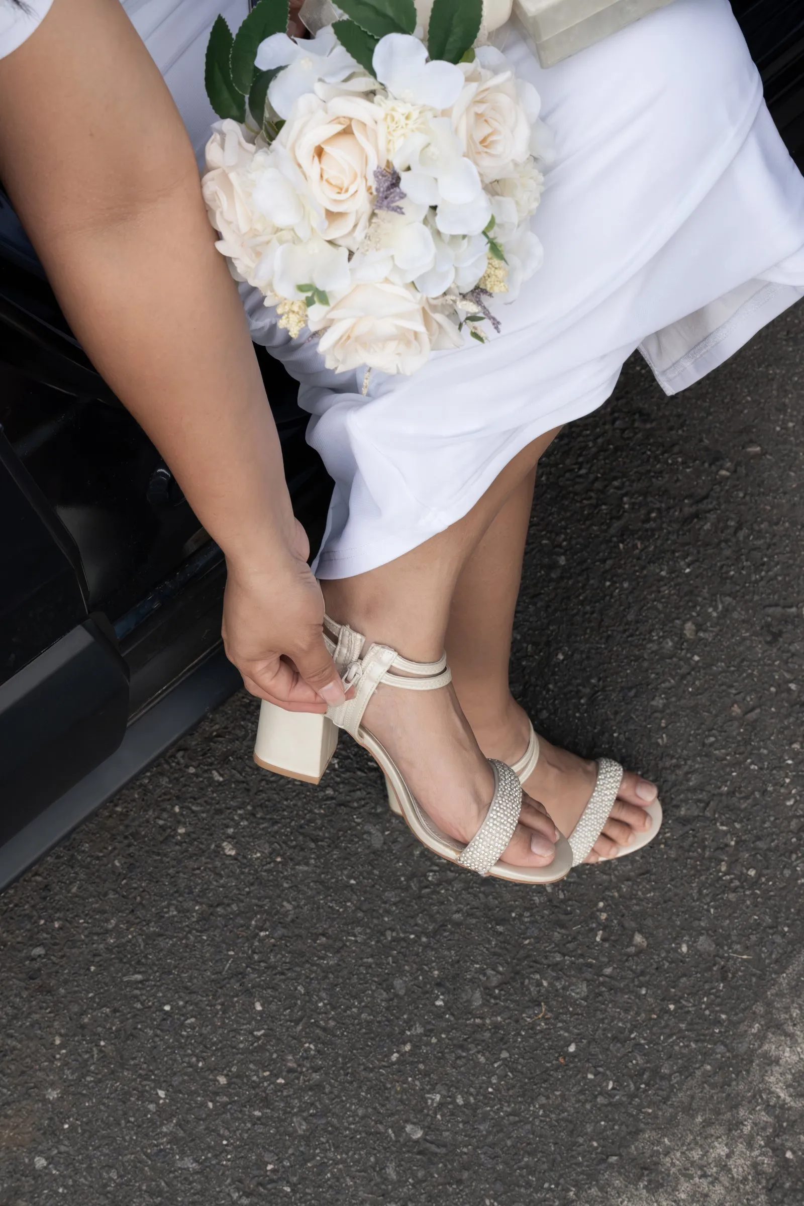 Winnie fastening the strap of her ivory block heel sandal, a bouquet of cream roses in her lap and her white gown draped to the pavement