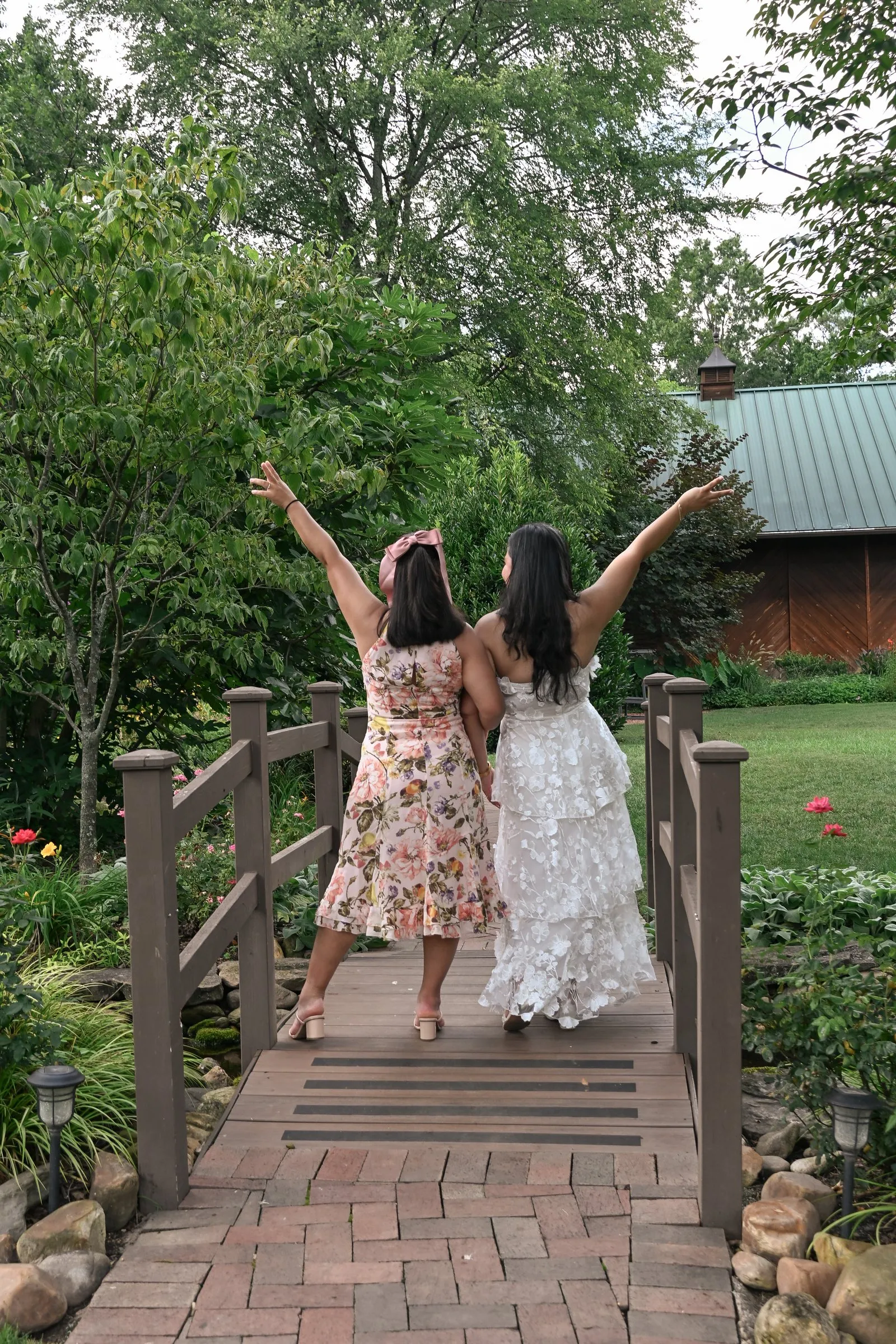 Winnie and her bridesmaid seen from behind with their arms raised on the footbridge, the red barn at Alexander Homestead beyond the trees
