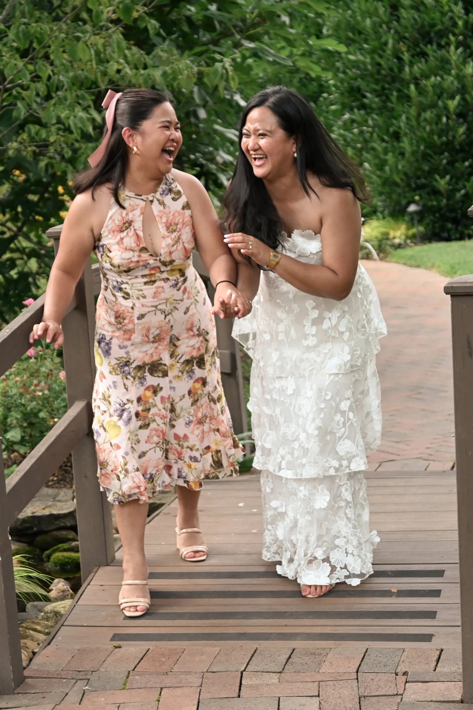 Winnie and a bridesmaid in a blush floral dress laughing as they cross the wooden garden footbridge at Alexander Homestead