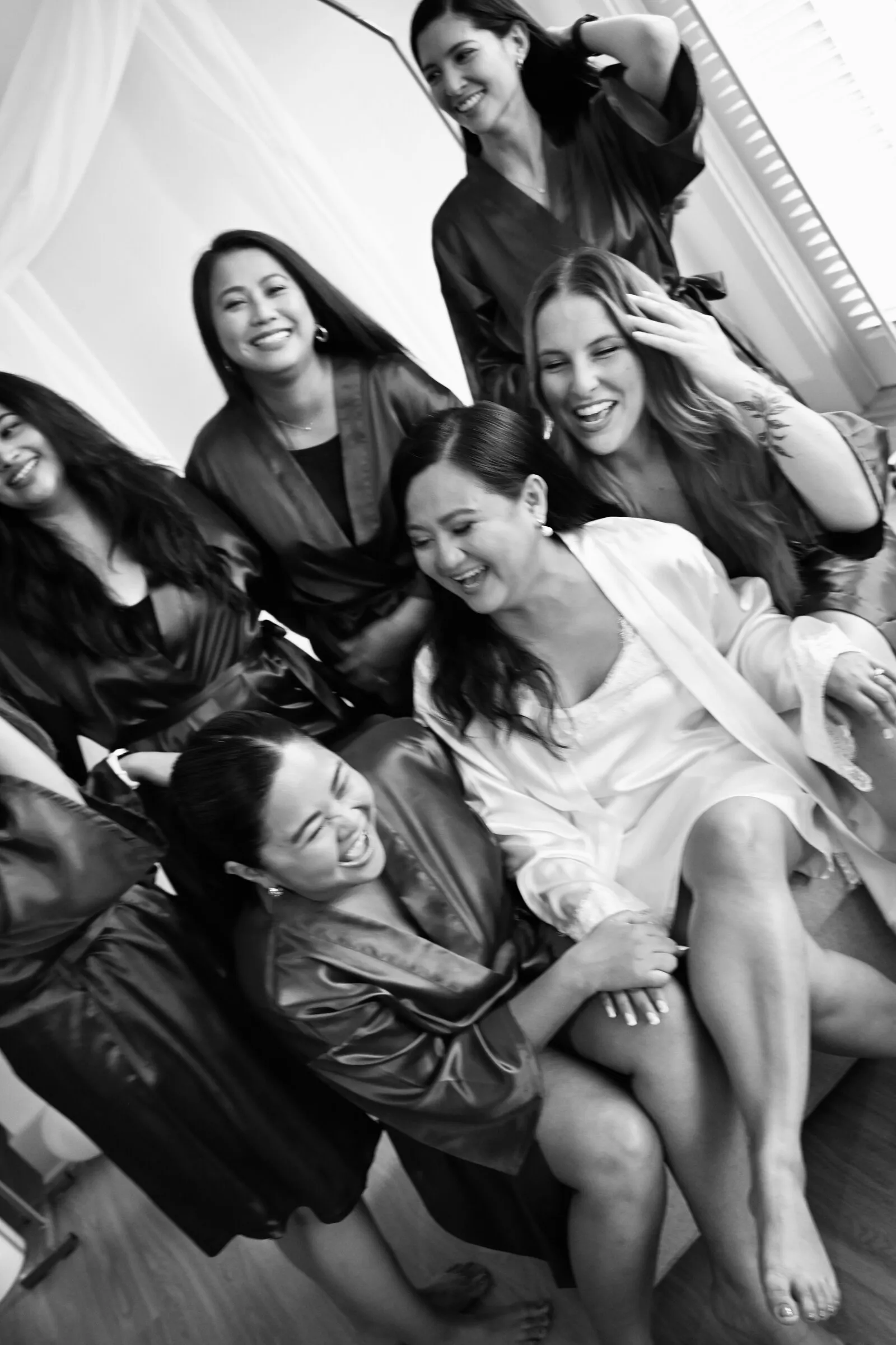 Black and white photograph of Winnie in her white robe laughing in a pile with her bridesmaids on a sofa, everyone cracking up at once