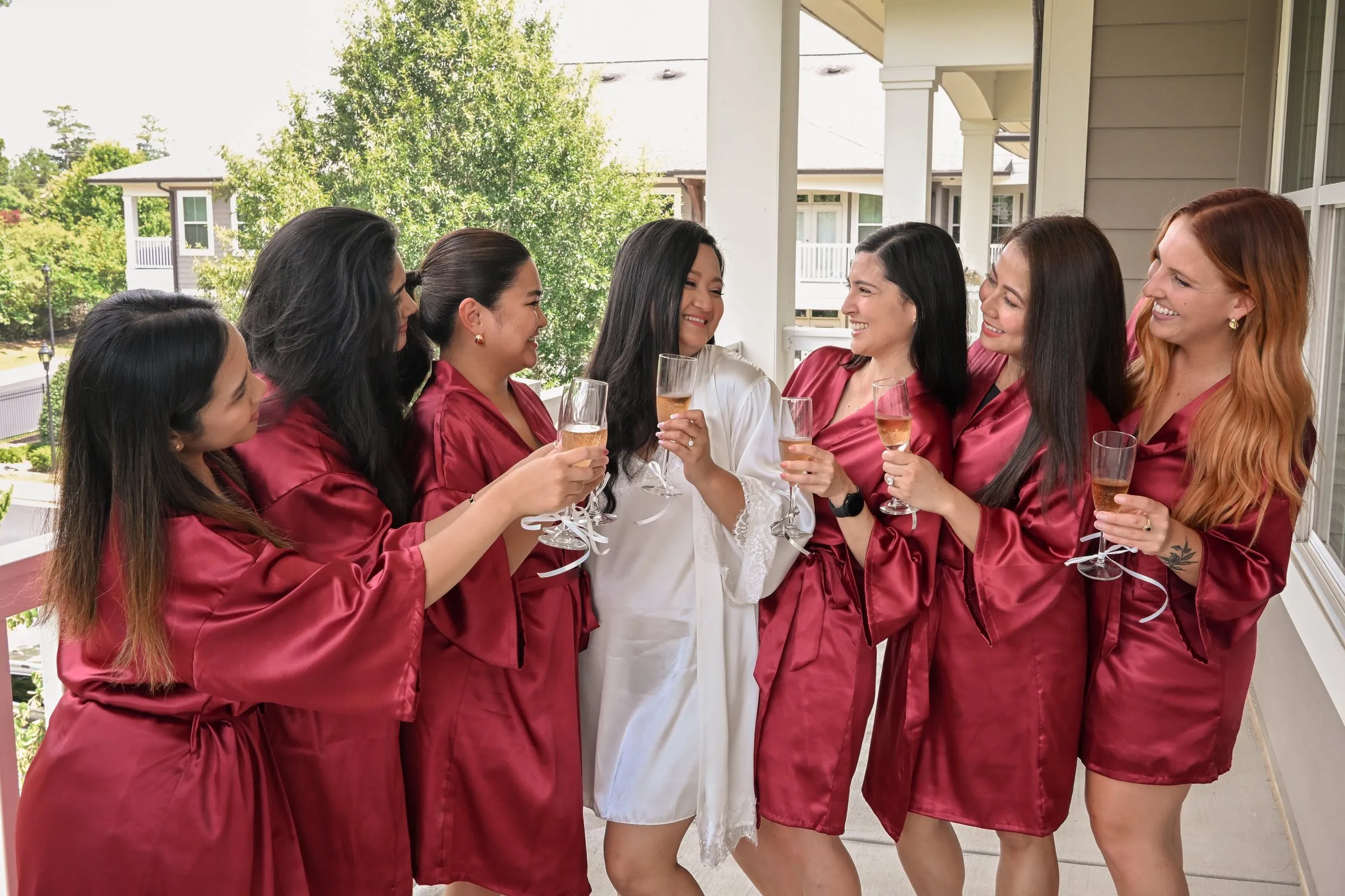 Winnie and her bridesmaids in red robes laughing and clinking champagne glasses on a covered balcony