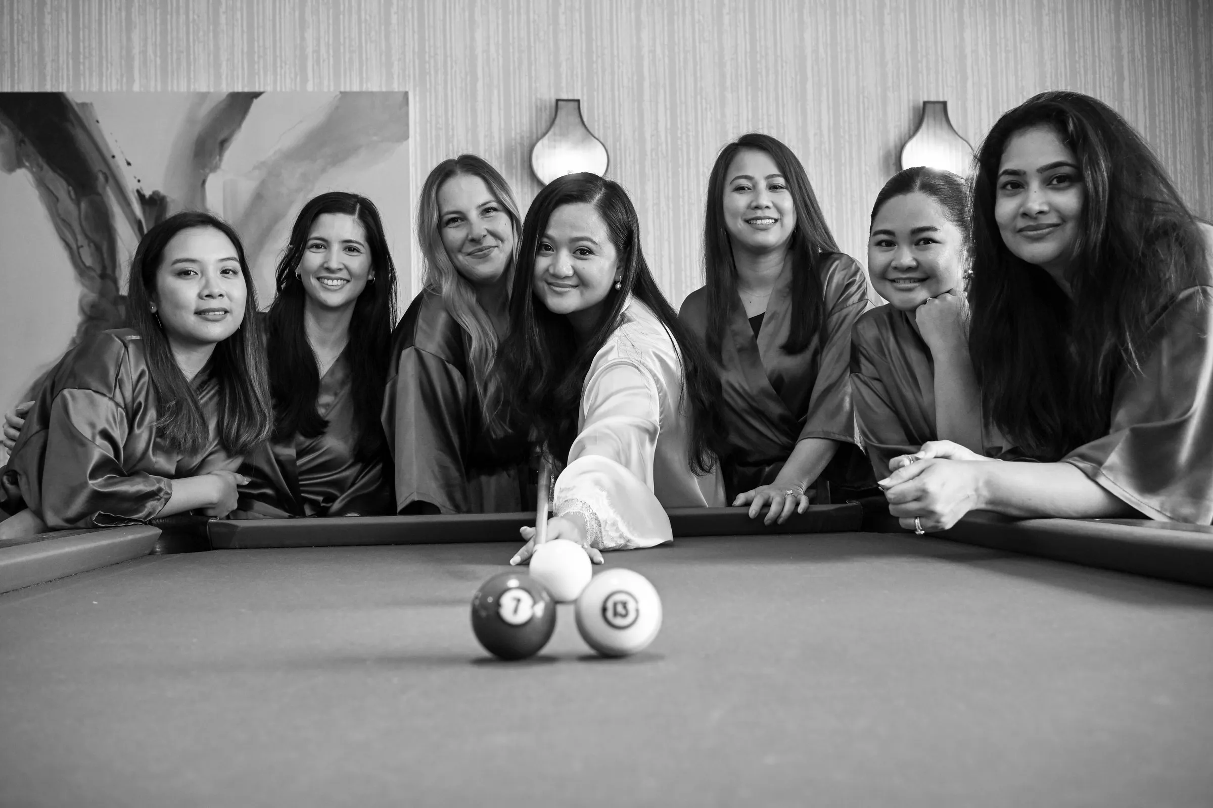 Black and white photograph of Winnie lining up a shot at the pool table with her bridesmaids behind her and the 7 and 13 balls set out for the wedding date