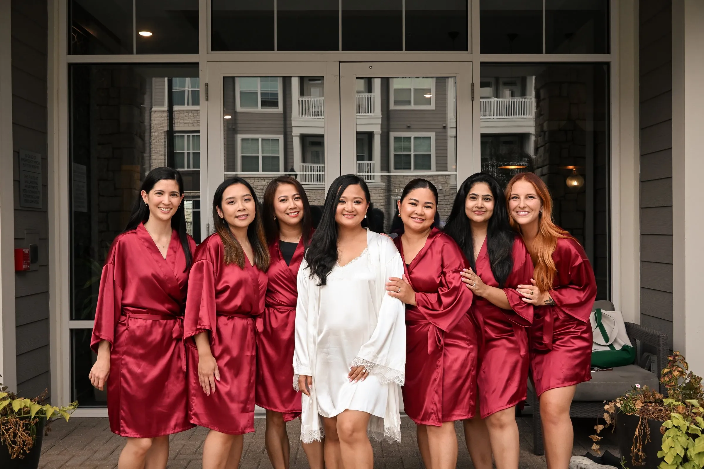 Winnie in a white robe standing with her bridesmaids in red satin robes outside a row of glass doors