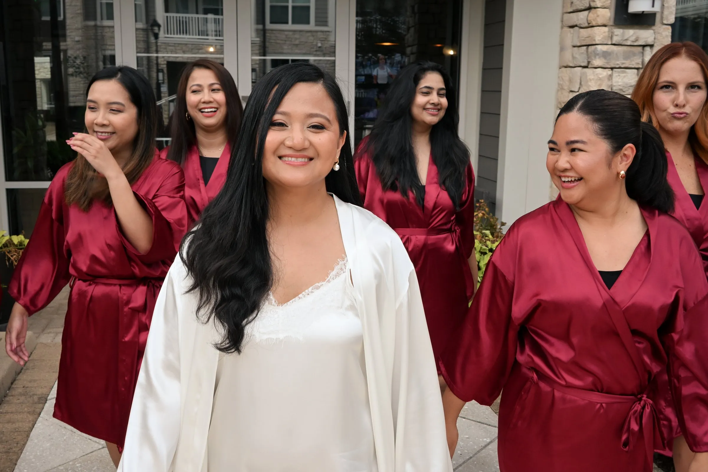 Winnie walking toward the camera smiling, her bridesmaids in red robes laughing behind her